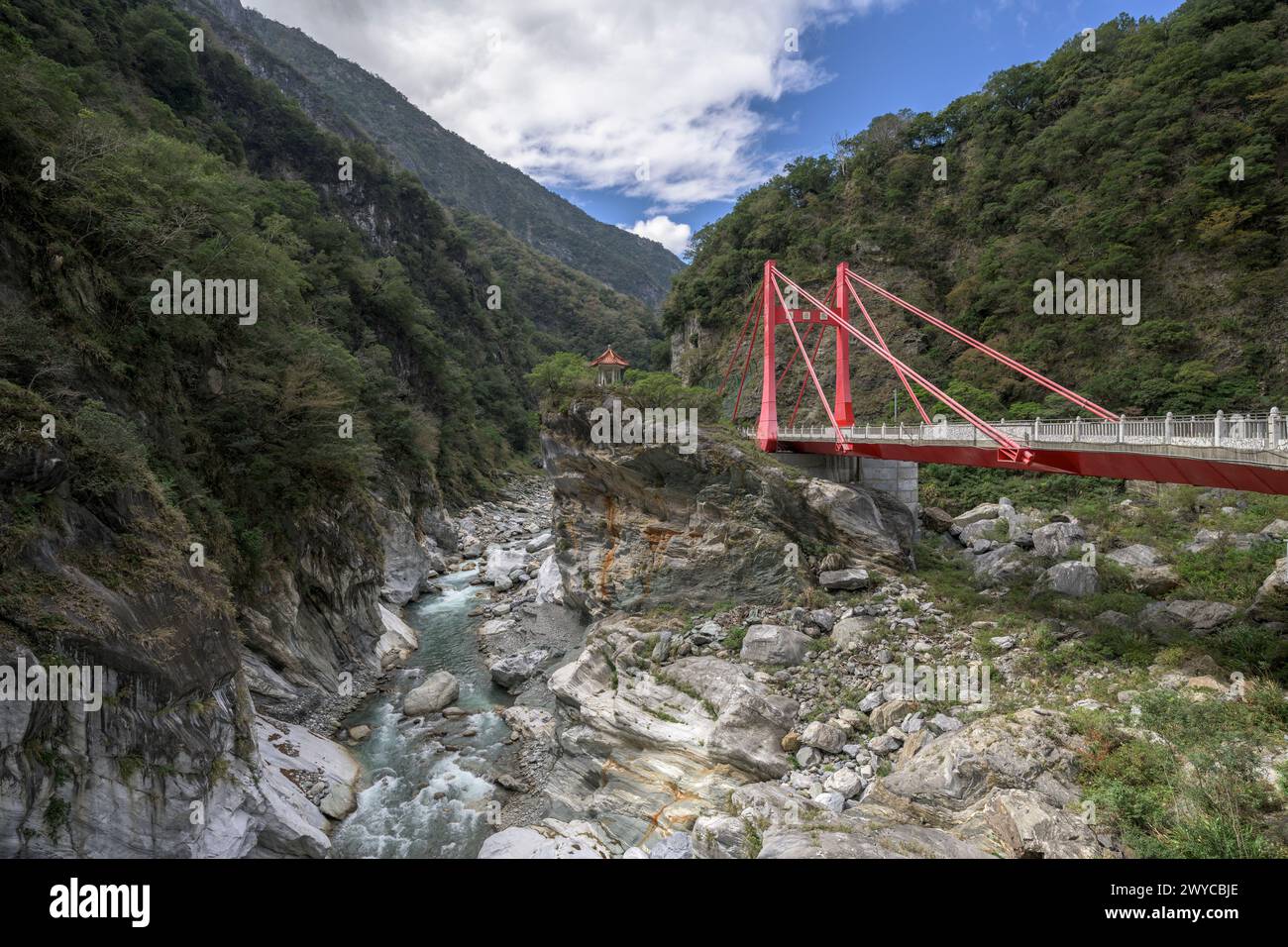 Un ponte sospeso rosso attraversa un fiume che attraversa la profonda gola rocciosa di Taroko circondata dal verde Foto Stock