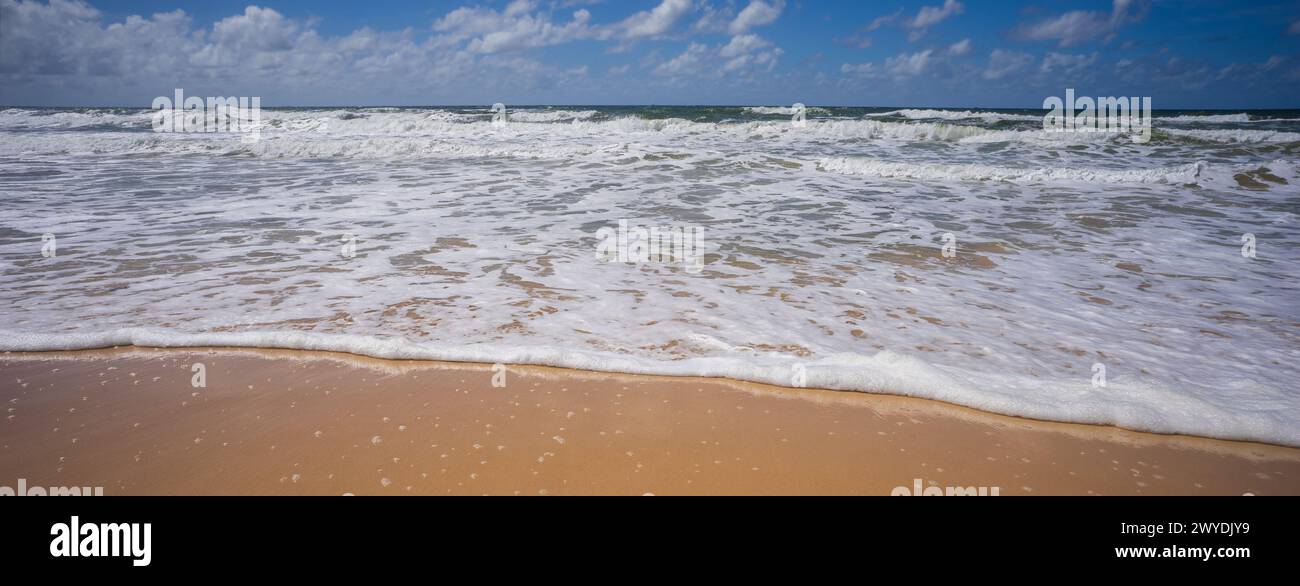 Una vista panoramica sull'oceano con onde rotanti, spiaggia di sabbia bagnata e cielo nuvoloso nei colori scuri del tardo pomeriggio Foto Stock