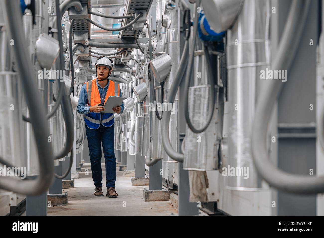 l'uomo in un giubbotto di sicurezza sta camminando attraverso un grande edificio industriale con tubi e fili. Ha un appunti che ispeziona l'area del gasdotto. Foto Stock