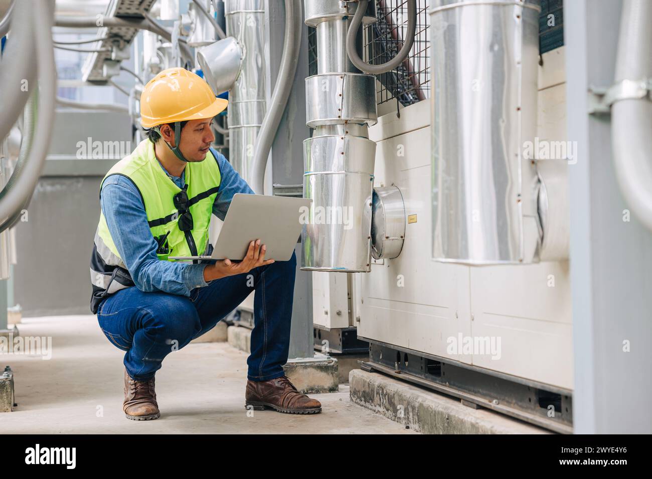 Un uomo con un giubbotto giallo guarda un notebook mentre si inginocchia. Indossa un elmetto e occhiali di sicurezza Foto Stock