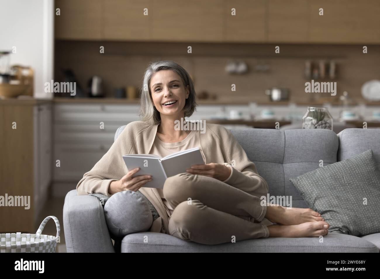 Donna di mezza età che posa per la macchina fotografica appoggiata sul divano con il libro Foto Stock