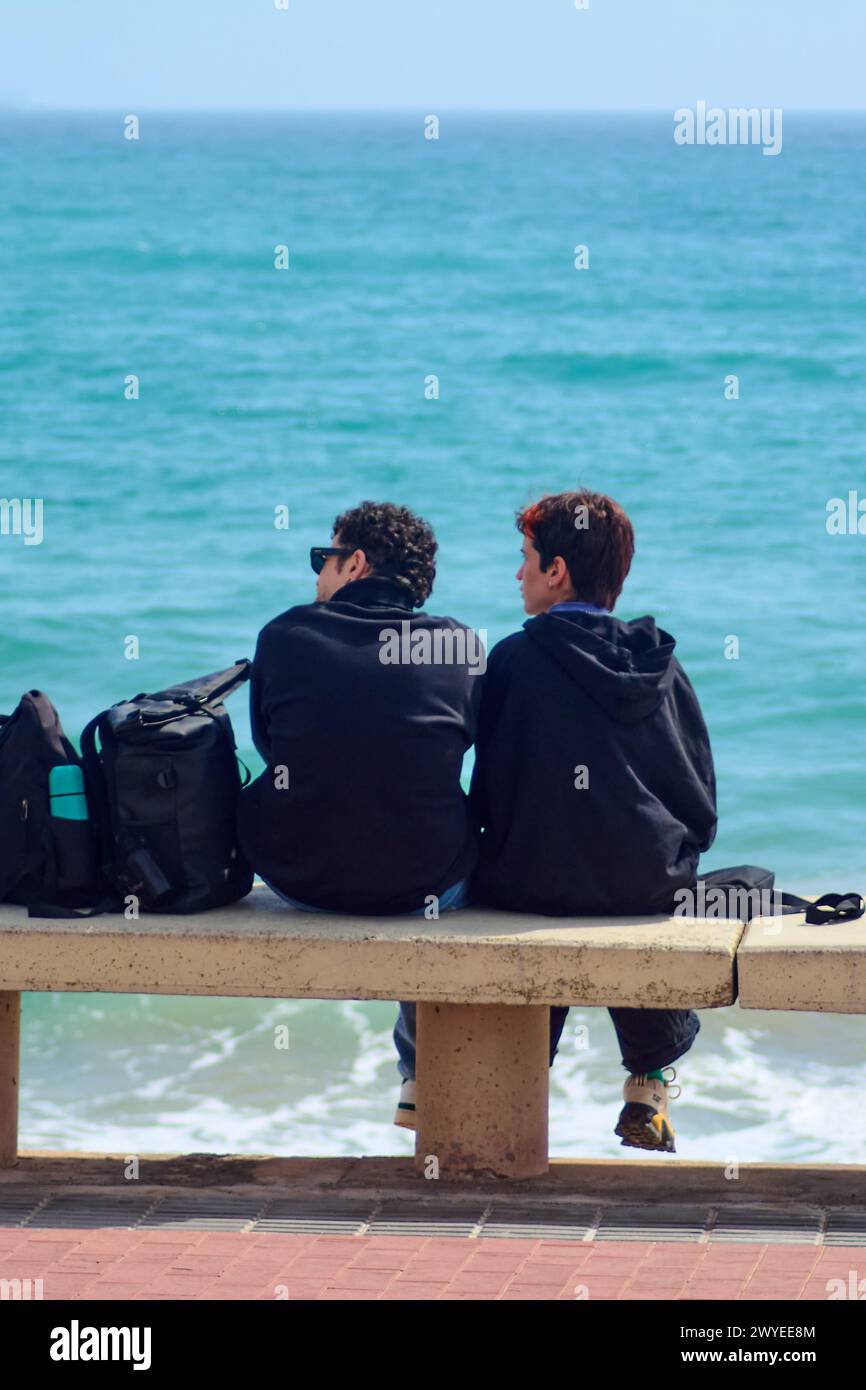 Tarragona, Spagna - 6 aprile 2024: Due amici condividono un momento di quiete seduto su un muro, con il vasto mare blu che si estende davanti a loro sotto una luce limpida Foto Stock