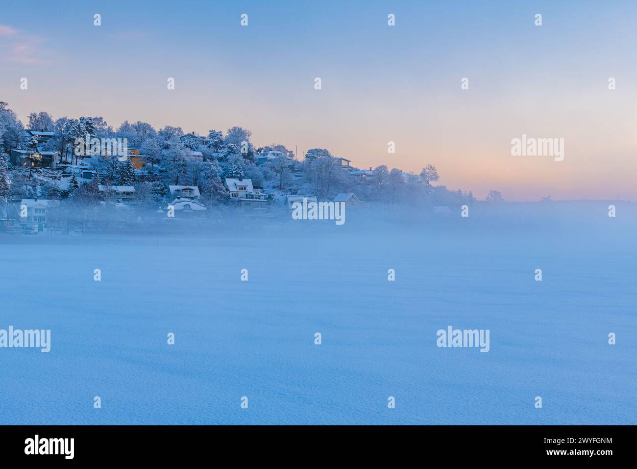 Un lago ghiacciato in primo piano con diverse case che punteggiano una collina in lontananza. Il paesaggio innevato è freddo e sereno, con la nebbia che aggiunge un Foto Stock