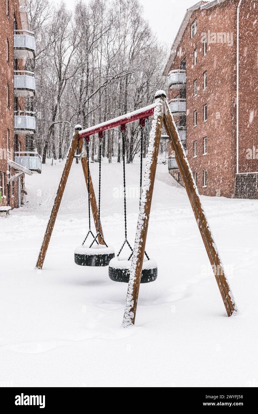 Un'altalena con sedili carichi di neve in posizione tranquilla in mezzo a una nevicata delicata. Lo sfondo presenta edifici residenziali in mattoni rossi, che incarnano un ser Foto Stock