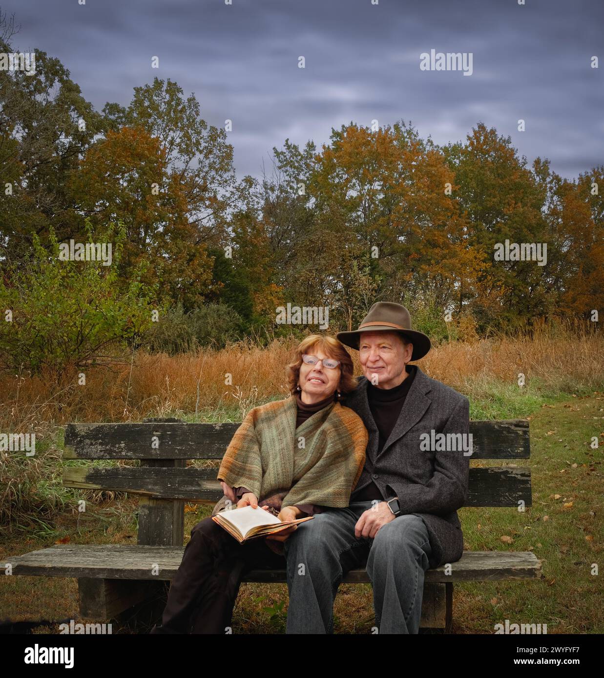 La coppia più anziana seduta sulla panchina nel parco il giorno coperto in autunno con una donna sorridente che tiene in mano un libro; l'uomo indossa un cappello largo Foto Stock