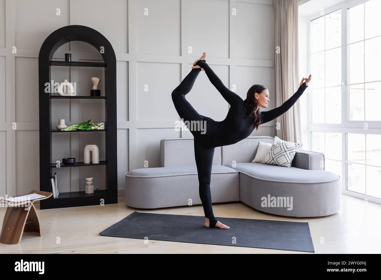 Attraente donna bruna che esegue l'esercizio Natarajasana a casa, danza in posa, pratica yoga in tute sportive nere in piedi su un tappetino, pieno Foto Stock