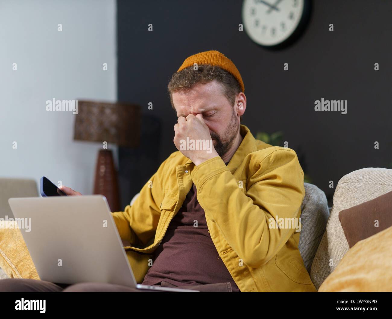 Un uomo con una giacca gialla è seduto su un divano con la testa tra le mani. Sta guardando un portatile ed è in stato di difficoltà Foto Stock