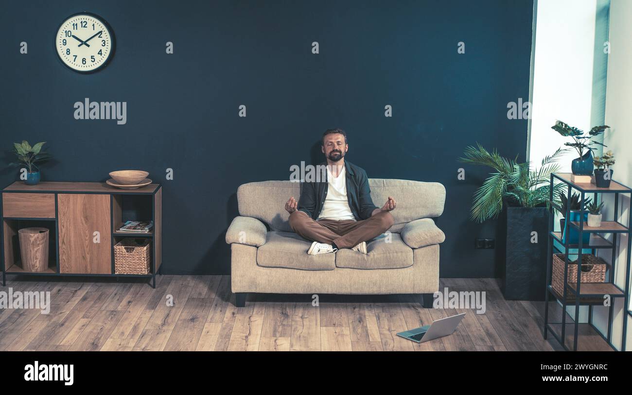 Un uomo è seduto su un divano in una stanza con un orologio sul muro. La camera è decorata con piante e un computer portatile è al piano terra. Quell'uomo è in relax Foto Stock
