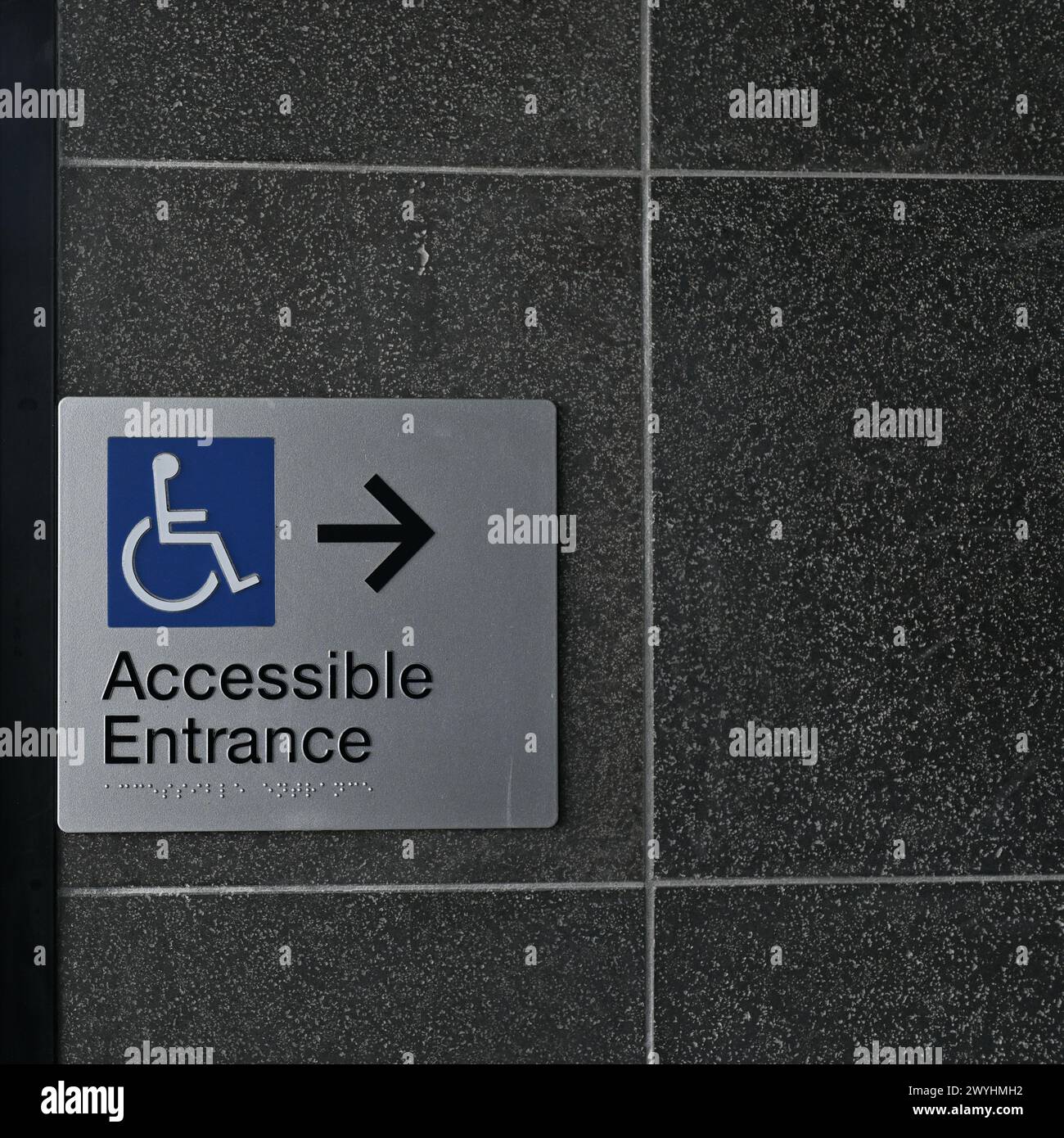 Cartello d'ingresso rettangolare accessibile con sedia a rotelle, con icona blu, testo nero, braille e una freccia nera su una parete scura Foto Stock