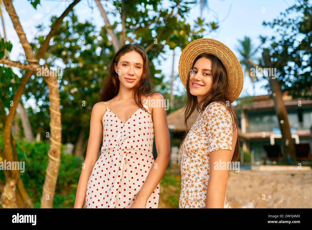 Due giovani donne con abiti vintage a pois e floreali si posano al resort tropicale sulla spiaggia, atmosfera estiva con stile retrò, amici sorridono vicino alle palme Foto Stock