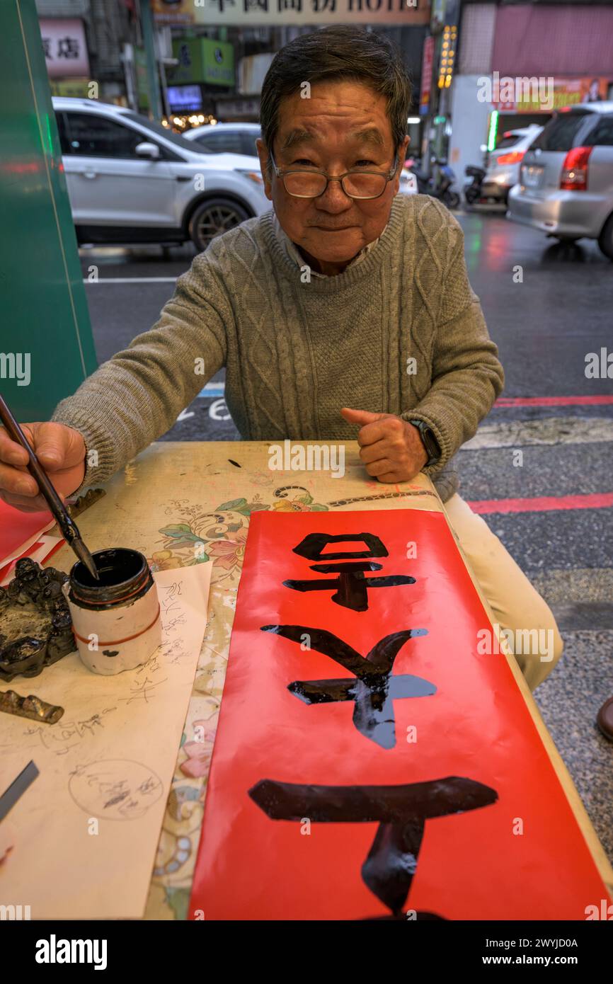 Un uomo anziano si concentra intensamente sulla scrittura di caratteri cinesi con un pennello su carta rossa, mostrando l'arte della calligrafia Foto Stock