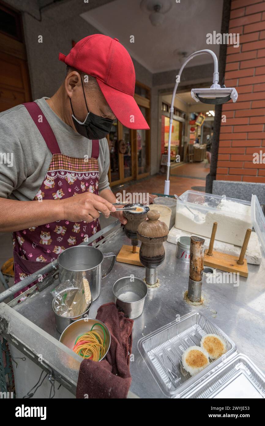 Un venditore ambulante che indossa una maschera prepara con attenzione i dolci tradizionali all'aperto utilizzando un utensile specializzato, durante il mercato cinese del capodanno Foto Stock