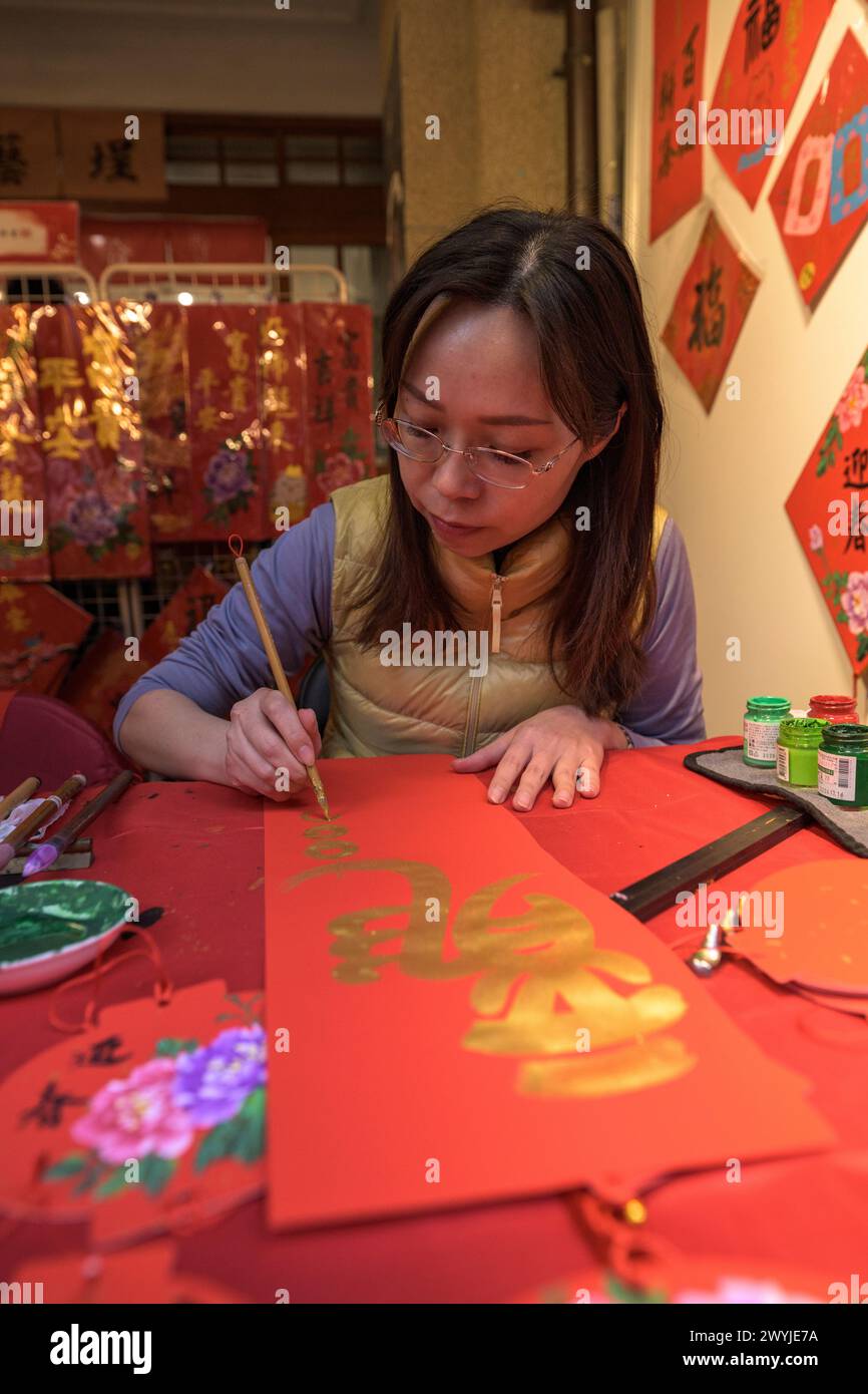 Una donna concentrata scrive calligrafia tradizionale cinese su carta rossa, facendo i desideri per il nuovo anno Foto Stock