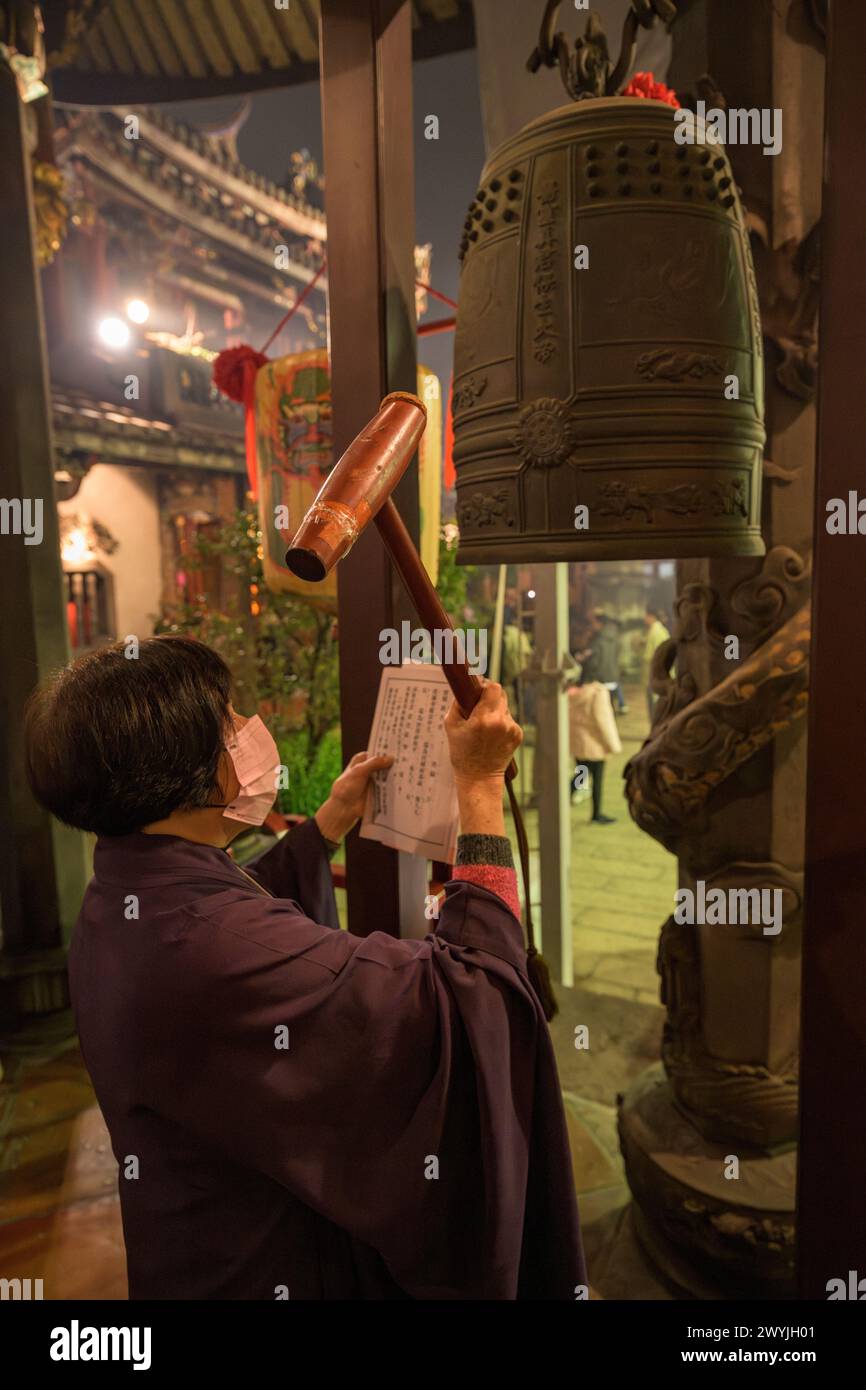 Donna abbigliamento tradizionale che suona una grande campana in una cerimonia del tempio Baoan, durante il capodanno cinese Foto Stock