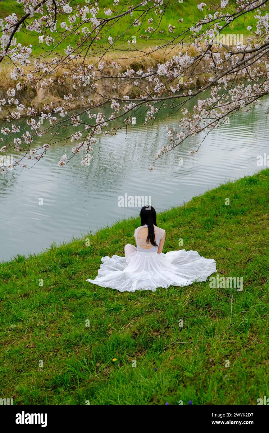 Persona in abito bianco seduta accanto a un fiume calmo sotto i fiori di ciliegio in primavera, catturando un momento tranquillo e riflessivo in Giappone. Foto Stock