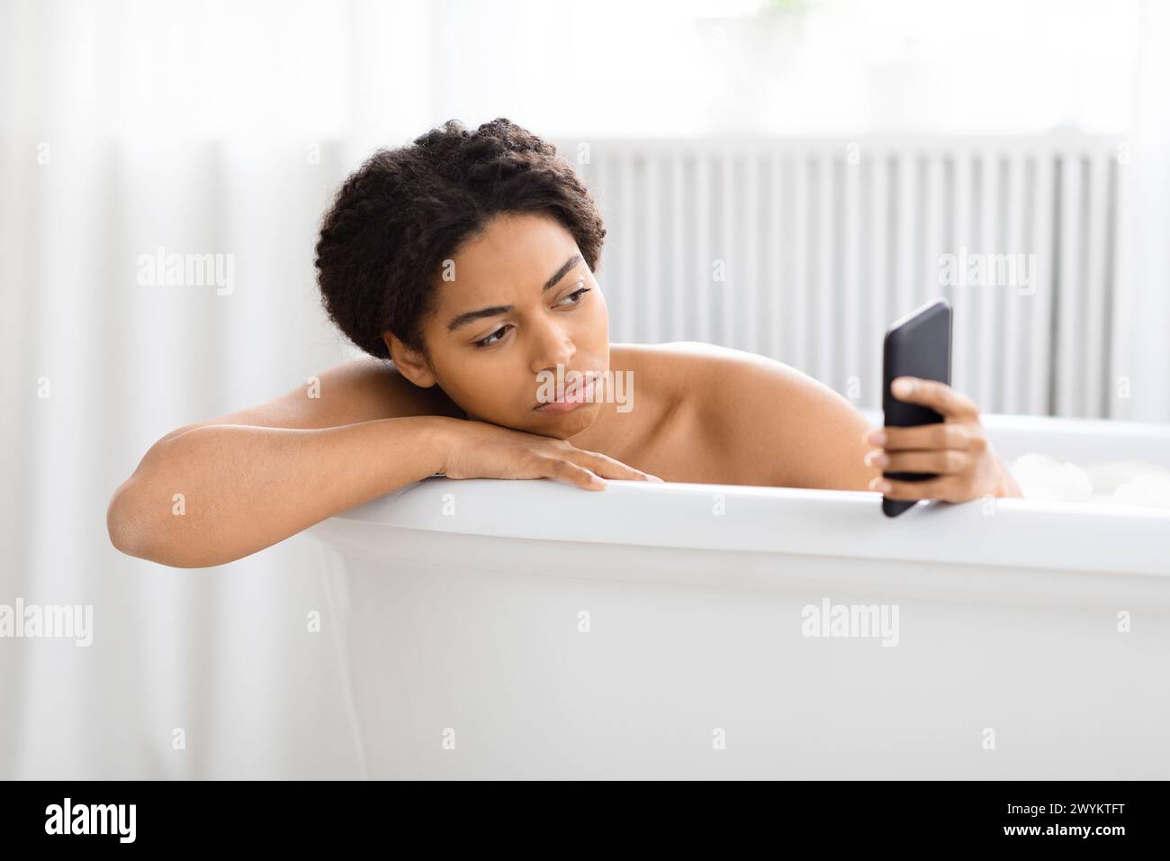 Pensiva donna afroamericana nella vasca da bagno con telefono Foto Stock