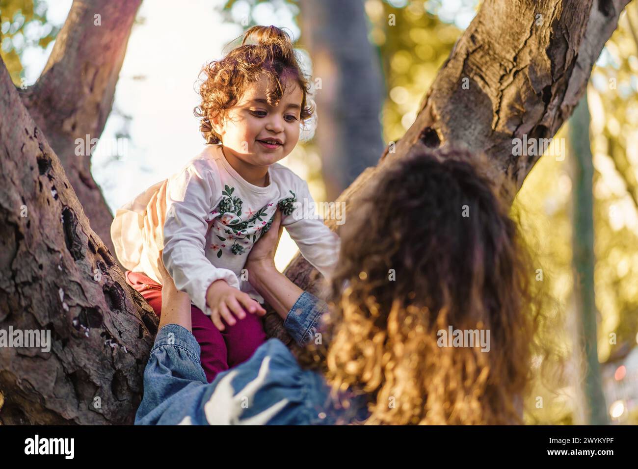 Nel cuore del parco, la cura di una mamma brilla mentre sostiene la scalata della sua bambina su un ramo d'albero: L'amore dei genitori e semplici gioie Foto Stock
