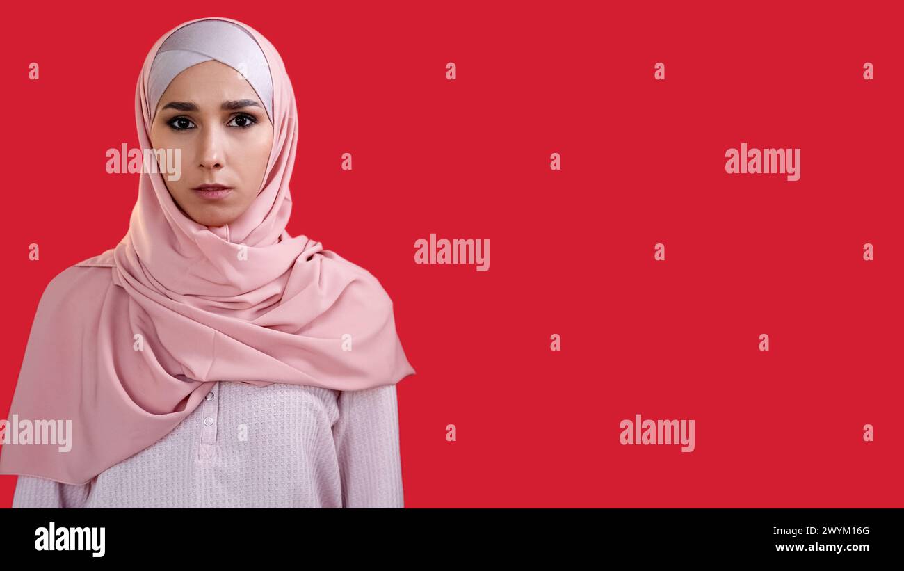 Ritratto femminile. Volto curioso. Bella donna sicura di sé con foulard hijab di colore rosa isolato su sfondo rosso vuoto. Foto Stock