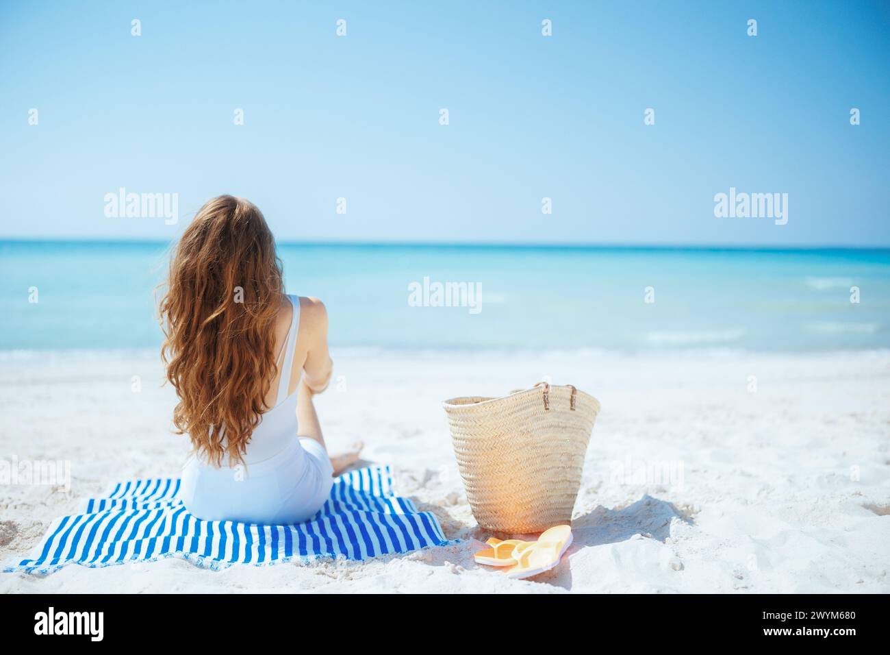 Vista da dietro elegante donna di 40 anni in riva al mare con un sacchetto di paglia e un asciugamano a righe. Foto Stock