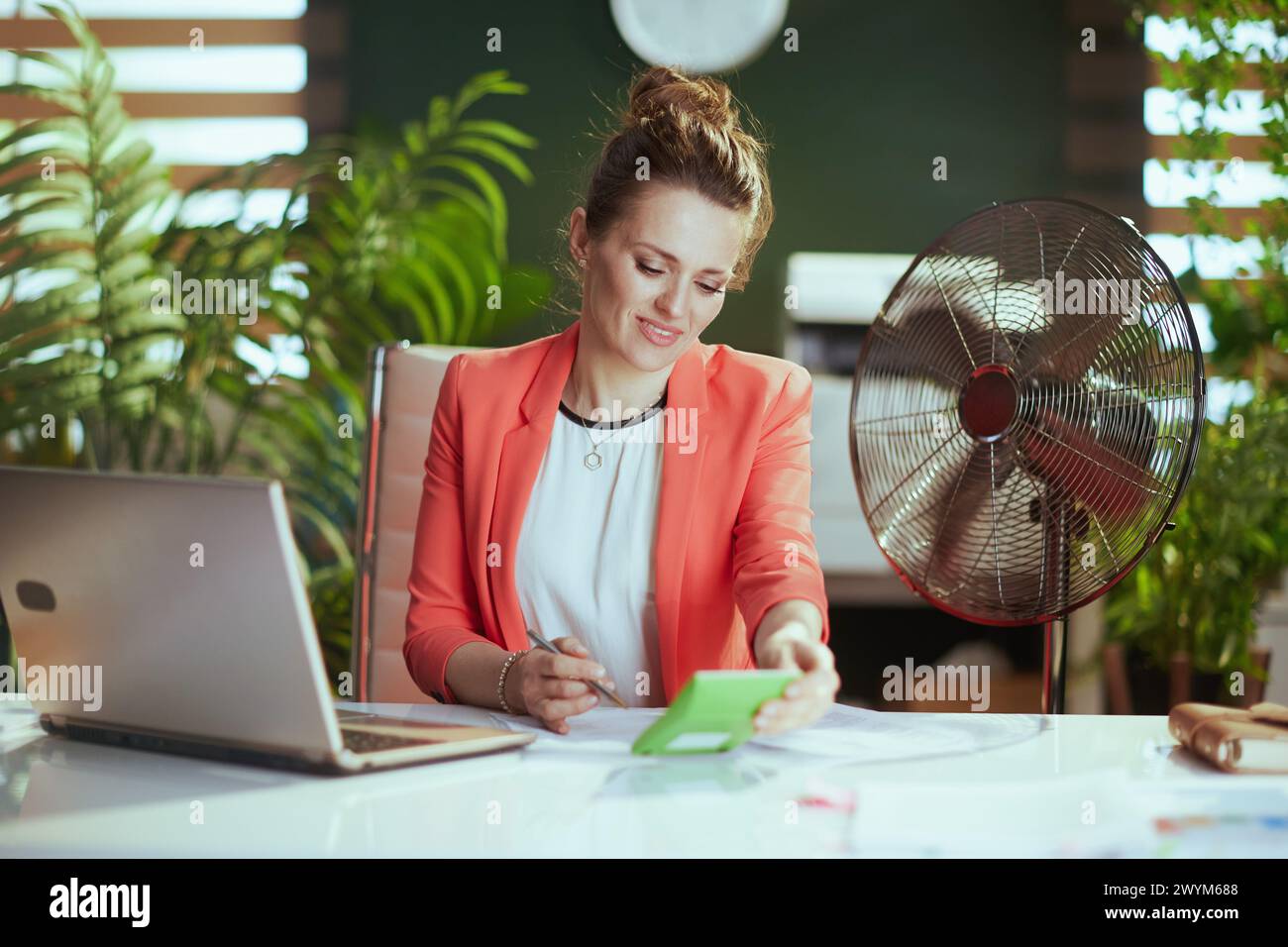 Ambiente di lavoro sostenibile. donna moderna e felice, al lavoro, con giacca rossa, calcolatrice, ventilatore elettrico e laptop. Foto Stock