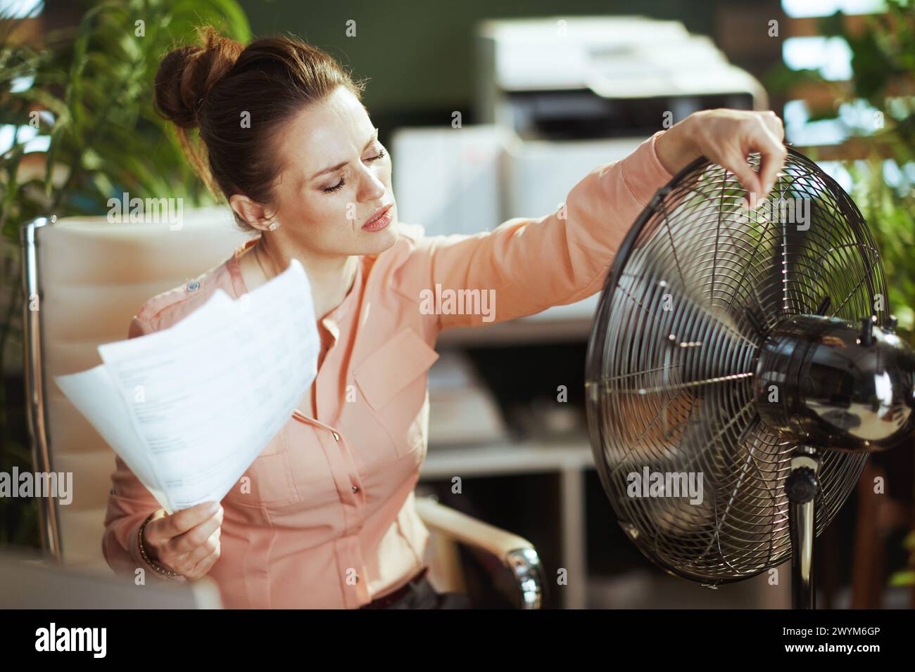 Ambiente di lavoro sostenibile. moderna dipendente donna di 40 anni in un moderno ufficio verde con documenti e ventilatore elettrico che soffrono di caldo estivo. Foto Stock