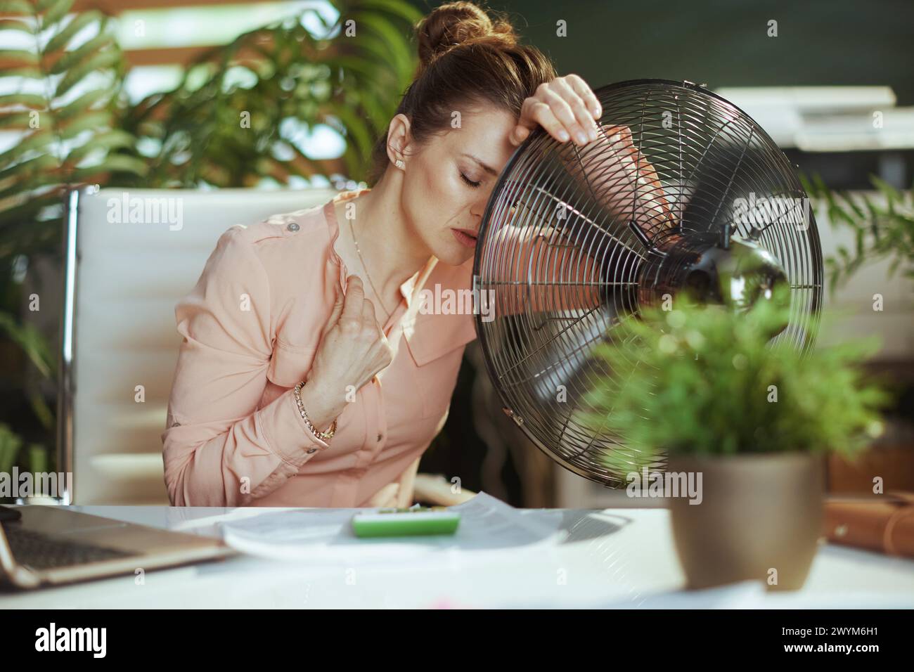 Ambiente di lavoro sostenibile. donna d'affari moderna di 40 anni al lavoro con ventilatore elettrico che soffre di caldo estivo. Foto Stock
