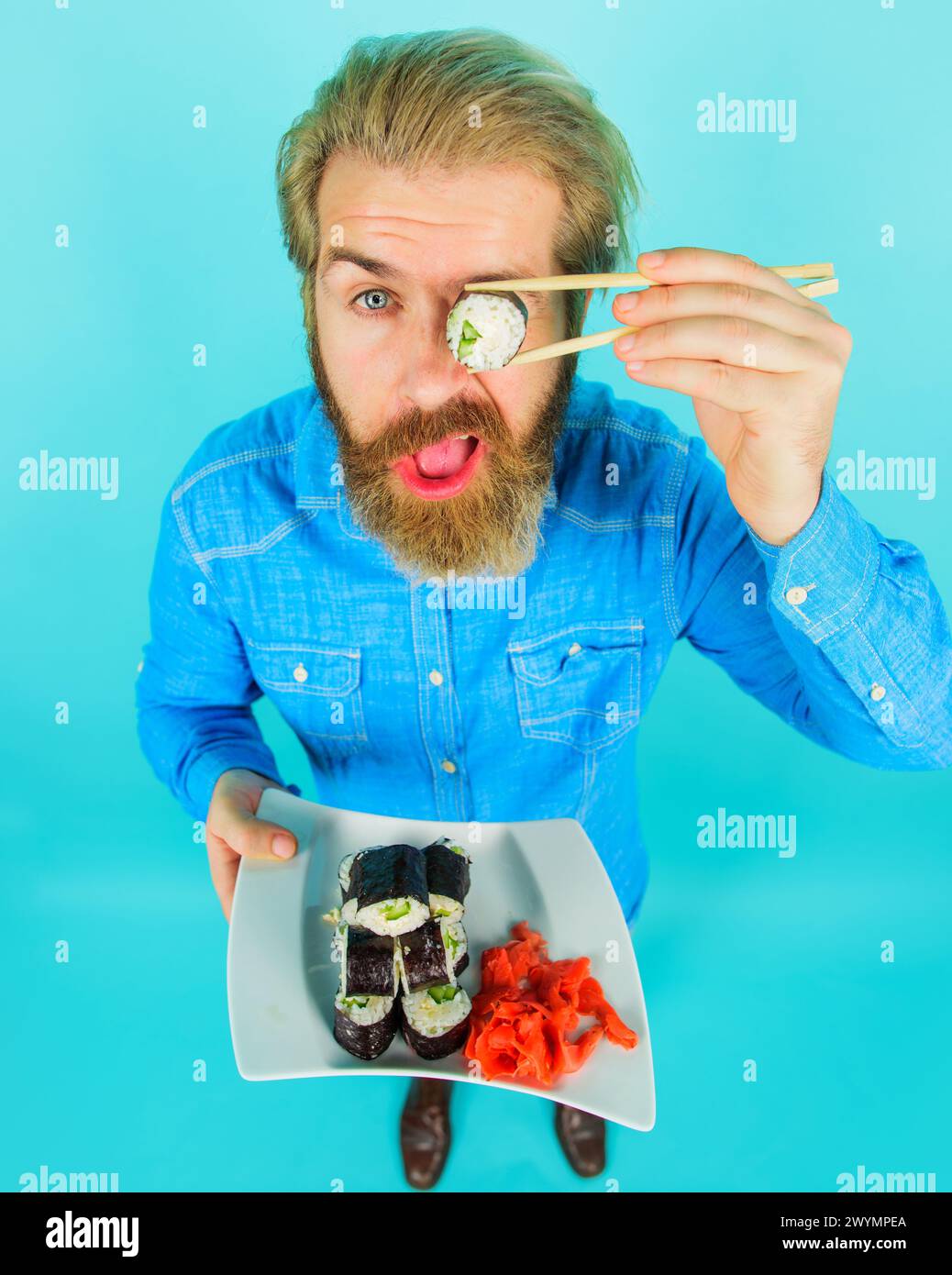 Consegna di sushi. Uomo con barba seria in denim casual che copre l'occhio con sushi roll. Cucina tradizionale giapponese. Bell'uomo con piatto di Foto Stock