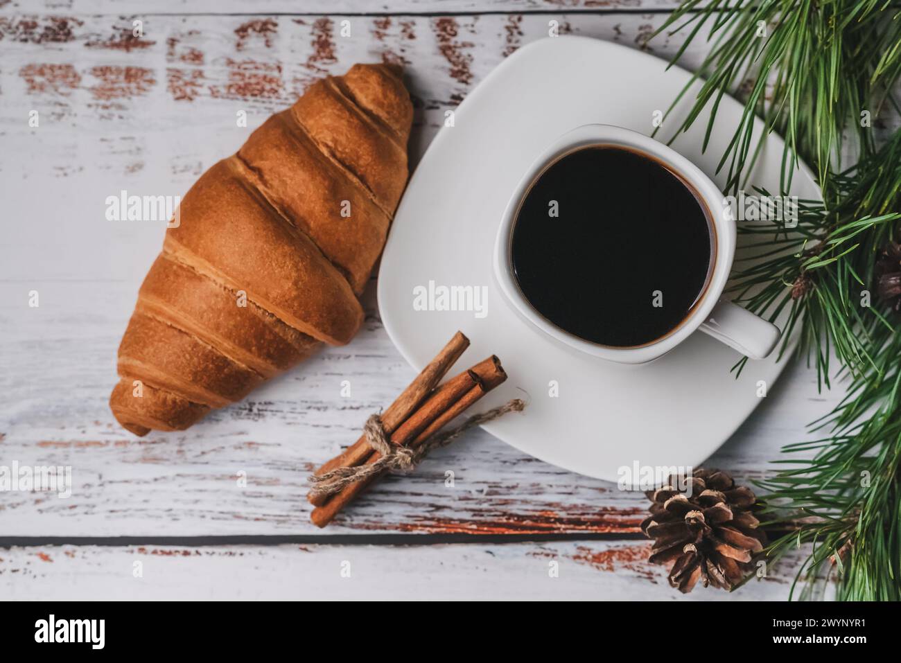 Colazione di Natale con una tazza di caffè, croissant e bastoncini di cannella. Tavolo chiaro in legno decorato con rami di abete e coni. Disposizione piatta, vista Foto Stock