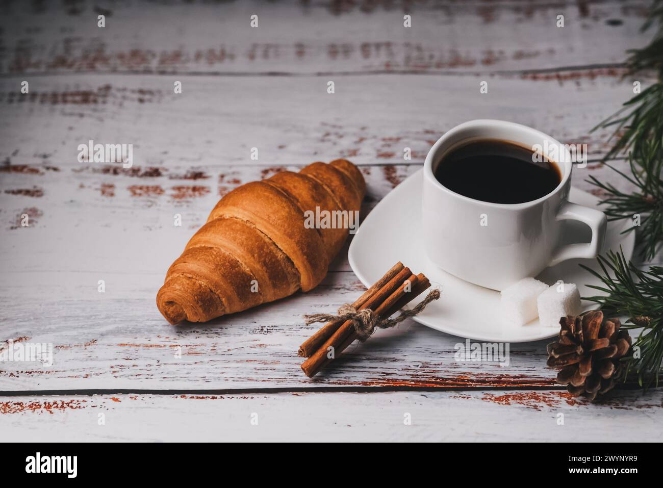 Colazione di Natale con una tazza di caffè, croissant e bastoncini di cannella. Tavolo chiaro in legno decorato con rami di abete e coni Foto Stock
