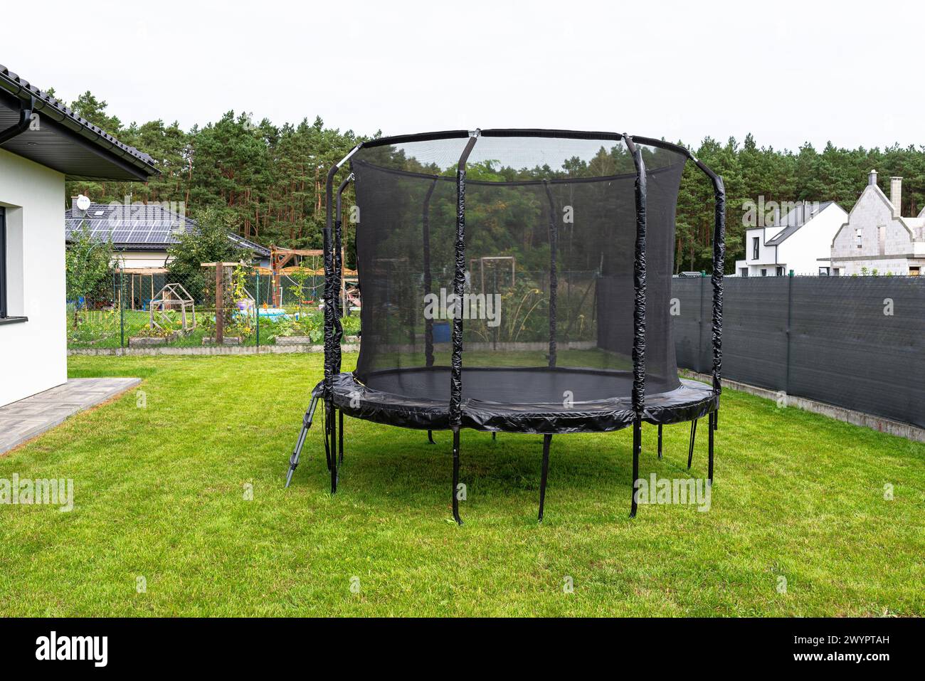 Trampolino per bambini di grandi dimensioni con rete protettiva e cerniera chiusa, in piedi in giardino, rete a vista che maschera la recinzione. Foto Stock