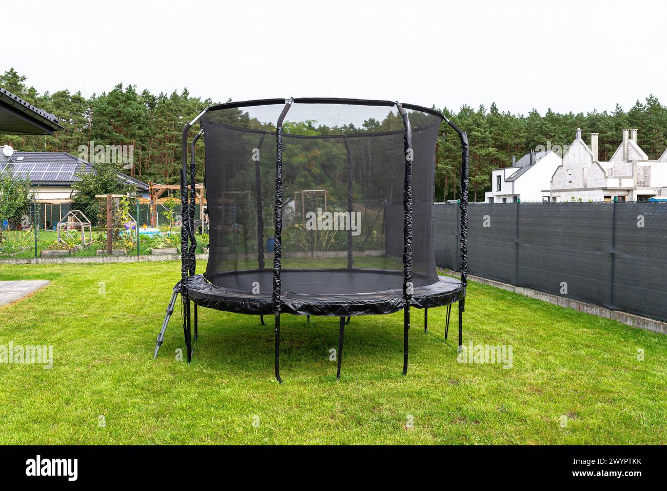 Trampolino per bambini di grandi dimensioni con rete protettiva e cerniera chiusa, in piedi in giardino, rete a vista che maschera la recinzione. Foto Stock