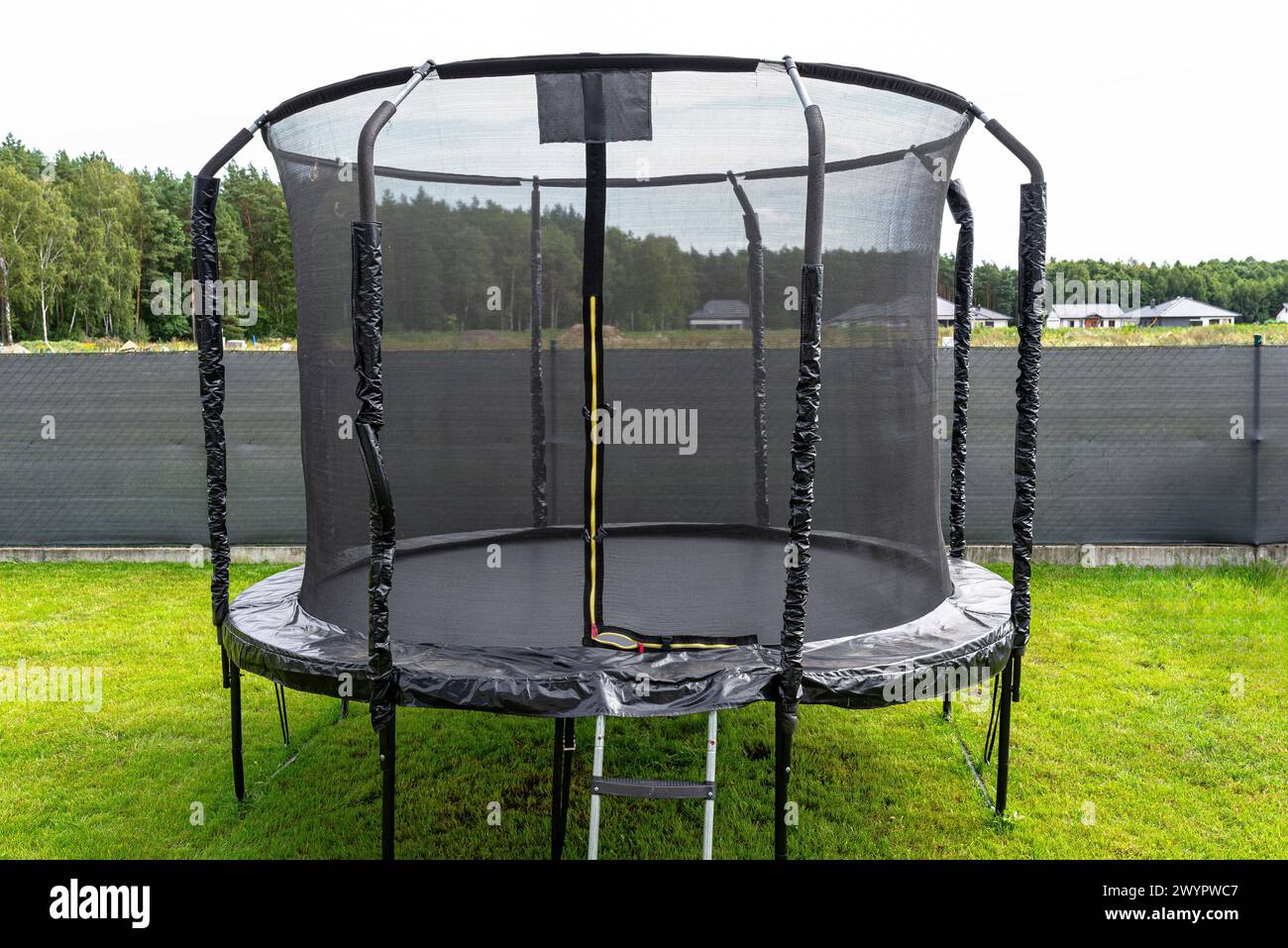 Trampolino per bambini di grandi dimensioni con rete protettiva e cerniera chiusa, in piedi in giardino, rete a vista che maschera la recinzione. Foto Stock