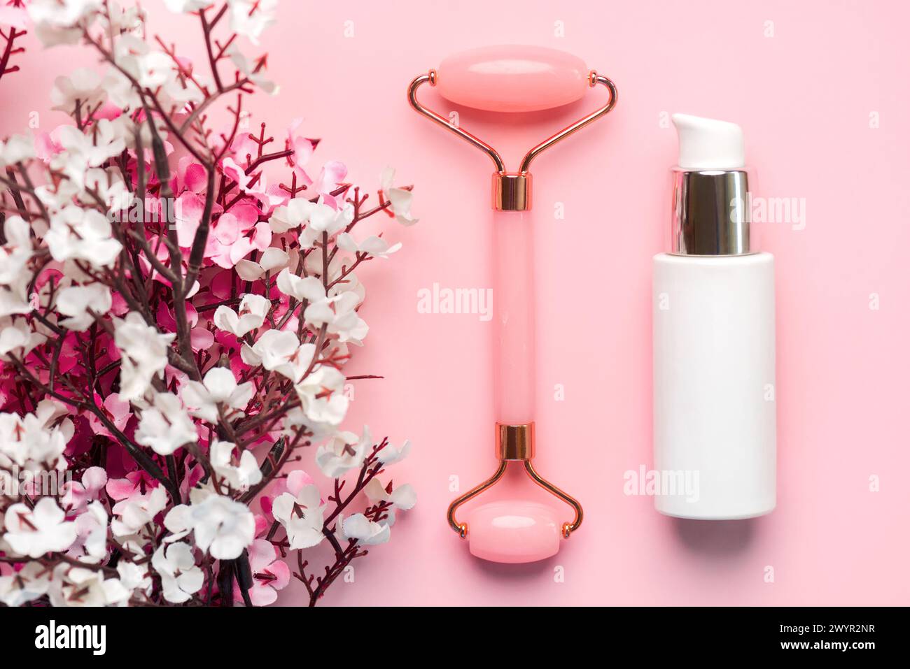 Primo piano del rullo massaggiatore di giada, bottiglia di crema viso per la cura della pelle e fiori di mandorle su sfondo rosa. Concetto di prodotti per la cura della pelle Foto Stock