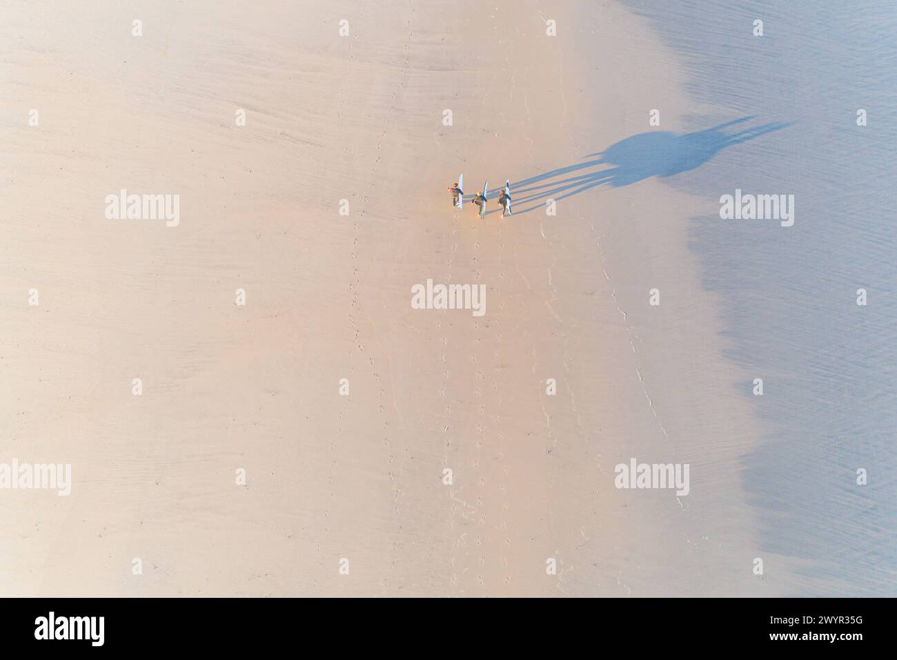 tre surfisti che portano le loro tavole da surf camminano lungo una spiaggia all'alba vista aerea con un drone Foto Stock