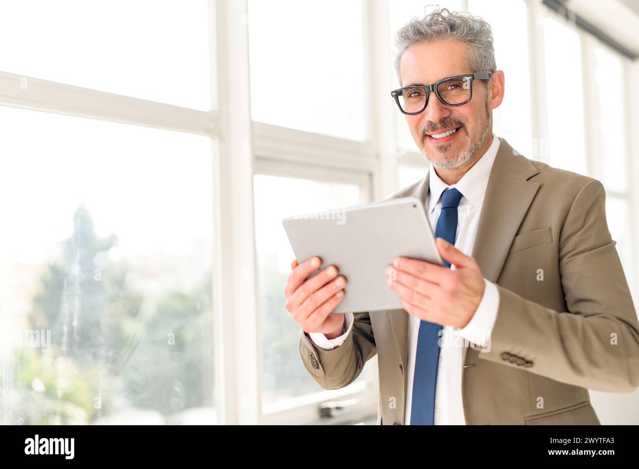 Un uomo d'affari senior si impegna con la tecnologia moderna, tenendo un tablet con un sorriso assicurato, mostrando come il tradizionale acume aziendale si fonde con gli strumenti moderni Foto Stock