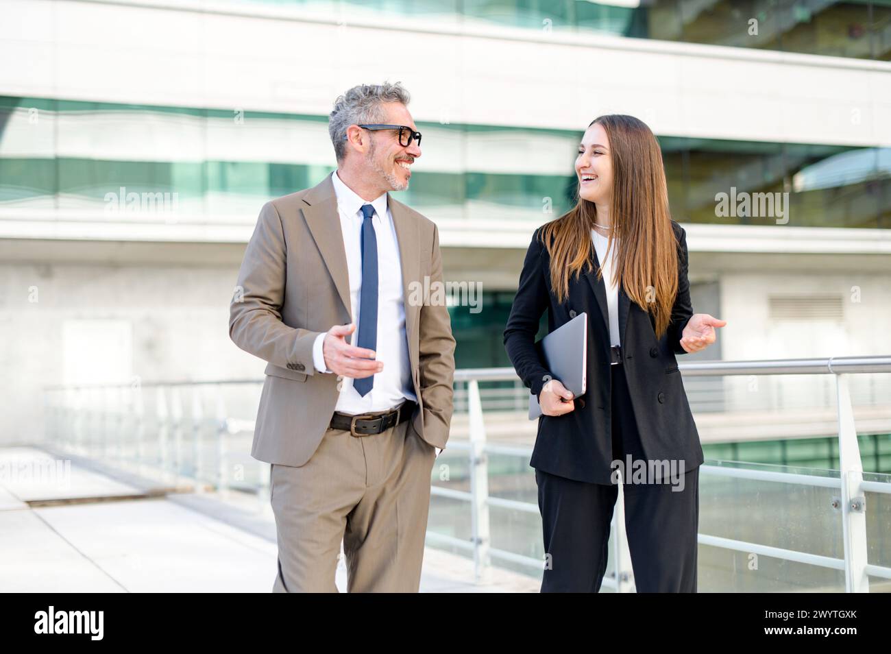 Un uomo d'affari maturo con i capelli grigi e un giovane collega sorridono mentre si impegnano in una discussione professionale contro l'ufficio moderno, evidenziando il valore della mentorship e della collaborazione nel business Foto Stock