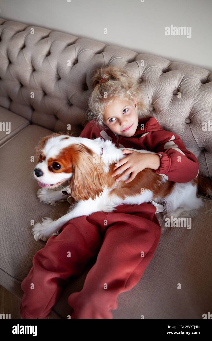 La bella bionda si siede a casa su un divano, giocando e abbracciando il cane, creando momenti emozionanti di gioia e compagnia con il cucciolo. Foto Stock