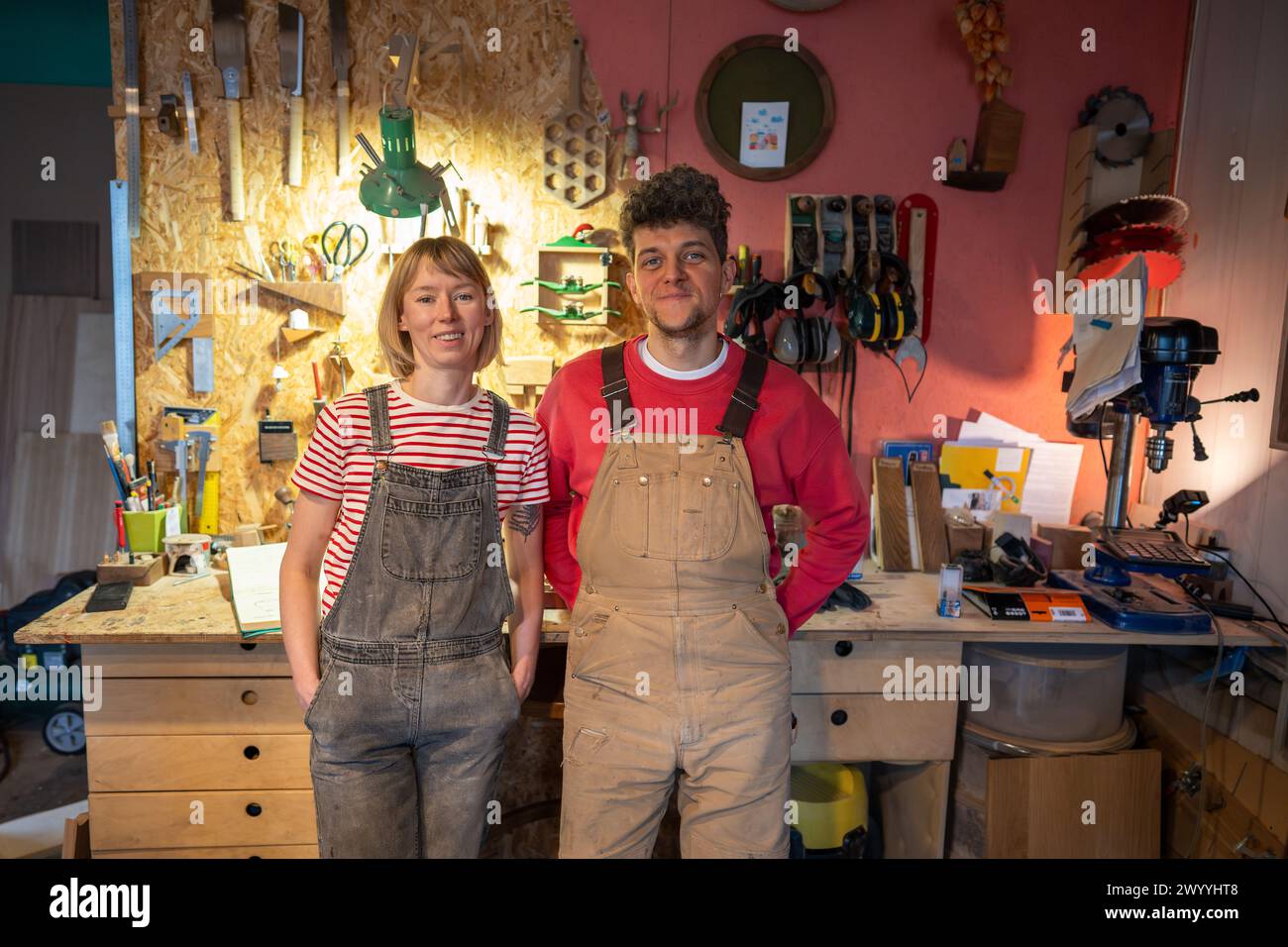 Una coppia di artigiani del legno che indossano tute in denim in piedi in una stanza artigiana insieme vicino al tavolo degli attrezzi Foto Stock