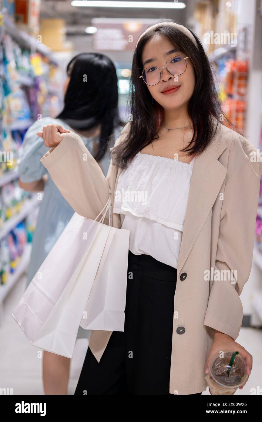 Una giovane donna asiatica sorridente si trova in un corridoio della spesa, tenendo borse della spesa, facendo shopping in un supermercato. concetti di shopping e stile di vita Foto Stock