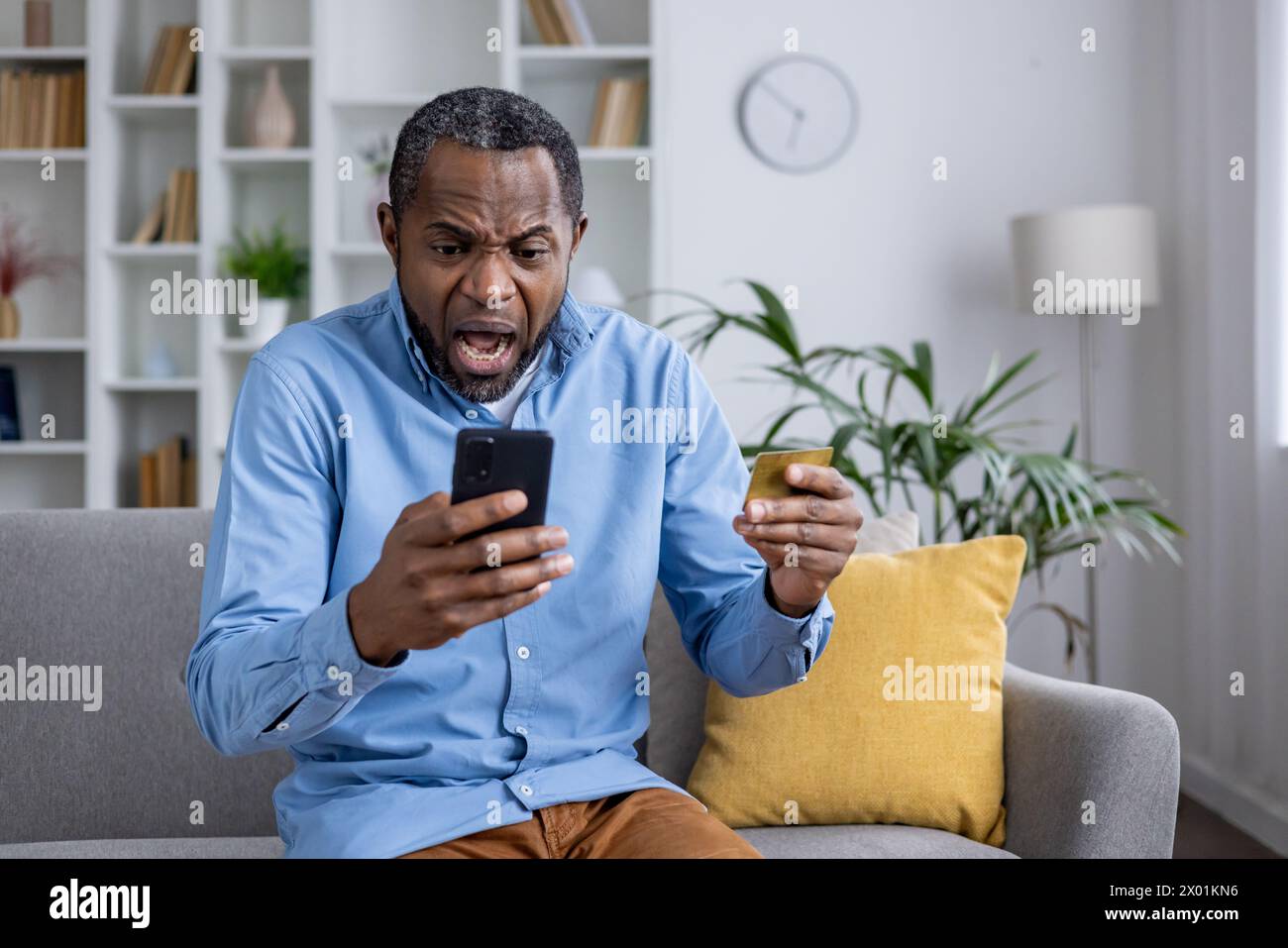 L'uomo seduto su un divano sembra scioccato e confuso, tenendo in mano una carta di credito e uno smartphone, realizzando attività fraudolente. Foto Stock