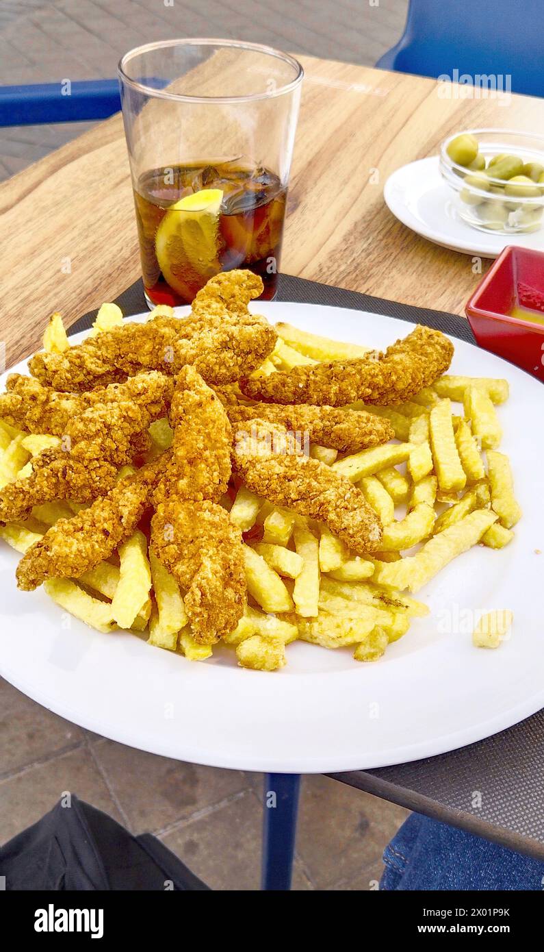 Un piatto di pollo fritto e patatine fritte si siede su un tavolo con un bicchiere di soda. Il piatto è pieno di cibo e la bibita è mezzo piena. La scena è casuale A. Foto Stock