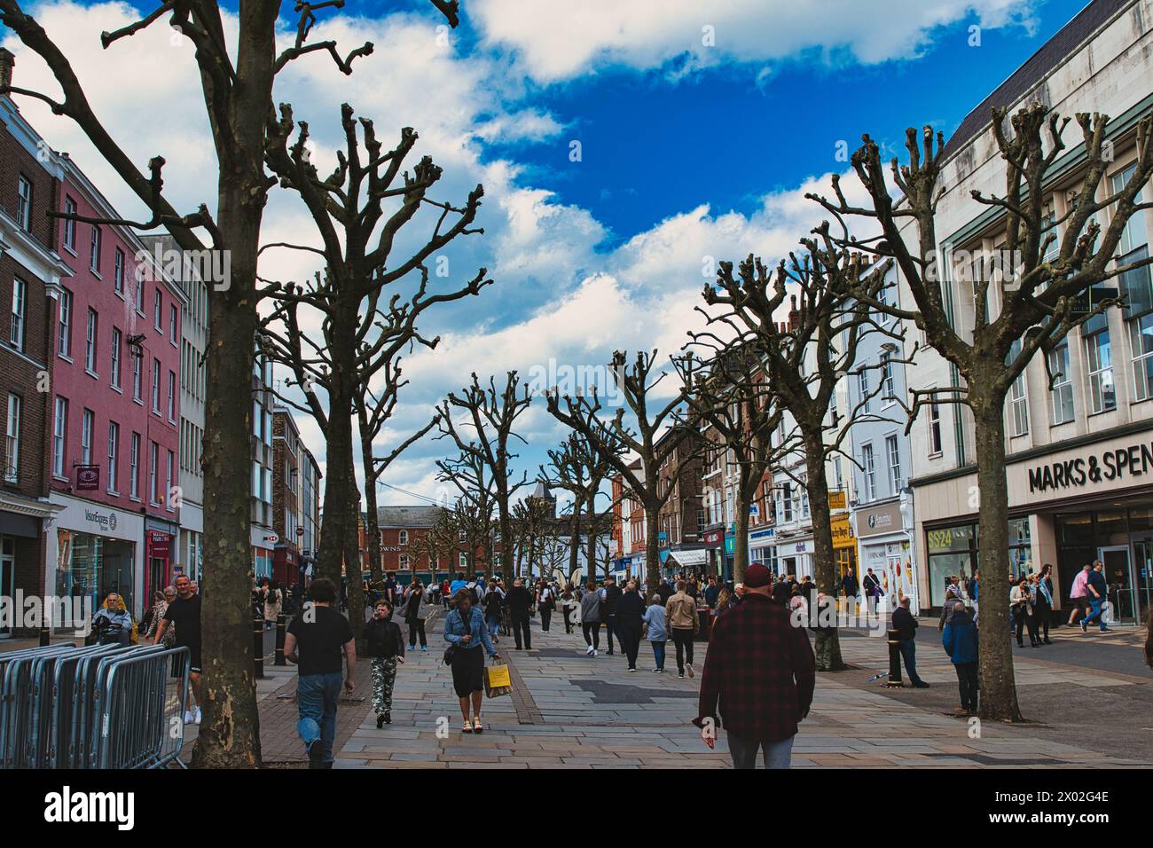 Vivace strada cittadina piena di pedoni, fiancheggiata da alberi potati senza foglie contro un cielo blu dinamico con soffici nuvole, che mostrano la vita urbana a. Foto Stock