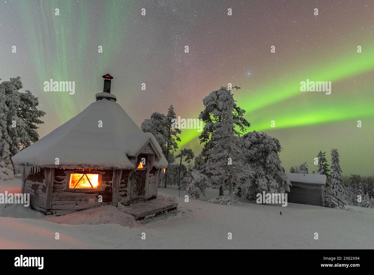 Sfumature di verde colorano l'aurora boreale (Aurora Boreale) sopra un paesaggio innevato con una tipica capanna illuminata da un fuoco all'interno in primo piano Foto Stock