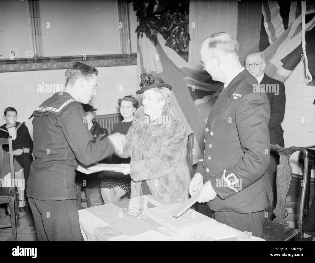 FESTA DI NATALE PER CADETTI DI MARE. 16 DICEMBRE 1944, AMMIRAGLIATO. FESTA DI NATALE DATA ALL'UNITÀ DELL'AMMIRAGLIATO DEI CADETTI DEL MARE NEL CLUB DEL PRANZO DELL'AMMIRAGLIATO. IL PRIMO ETERNO, IL SIGNOR A V ALESSANDRO, SI RIVOLSE AI CADETTI E LA SIGNORA ALESSANDRO PRESENTÒ I PREMI. - La sig.ra Alexander presenta il "Quiz" primo premio per la marineria all'agente Richard Fairbairn, di Grays Essex, Libro di Godfrey Winn "Home from Sea" Foto Stock