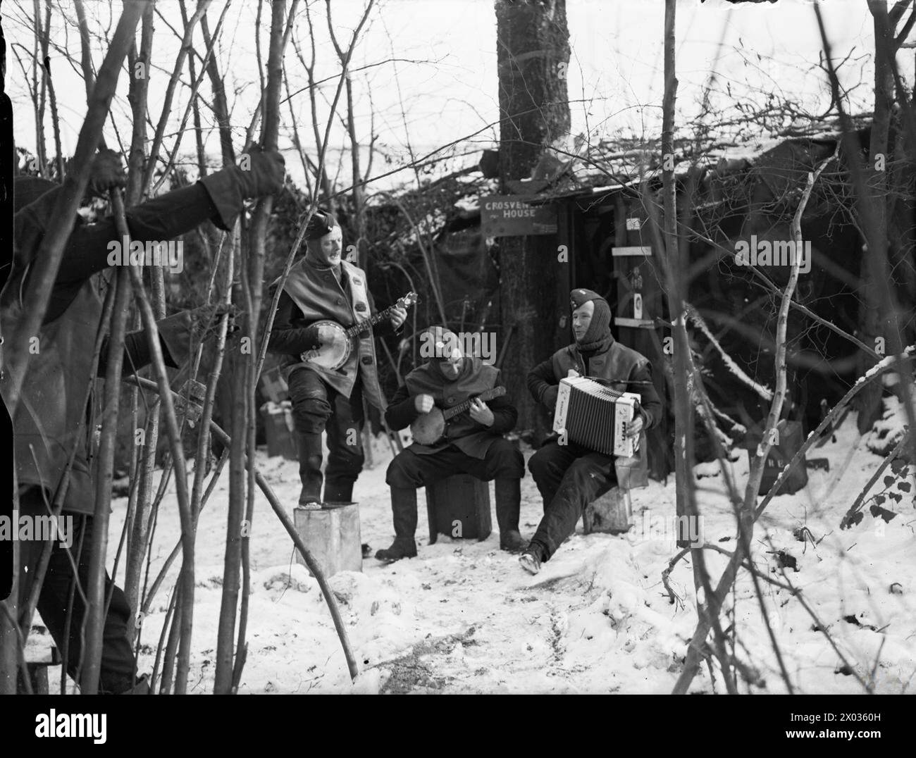 Tre aviatori della RAF della "Grosvenor House Orchestra" si esibiscono con un ukulele e un melodeon per intrattenere gli spettatori fuori dal loro billet su un campo d'aviazione coperto di neve in Francia, 1939-1940. Foto Stock Tre aviatori della RAF della "Grosvenor House Orchestra" si esibiscono con un ukulele e un melodeon per intrattenere gli spettatori fuori dal loro billet su un campo d'aviazione coperto di neve in Francia, 1939-1940. Foto Stock