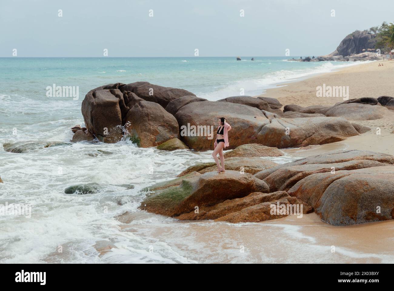 La donna passeggia lungo una costa punteggiata di massi, con onde che si infrangono dolcemente e mare turchese che si estende fino all'orizzonte Foto Stock