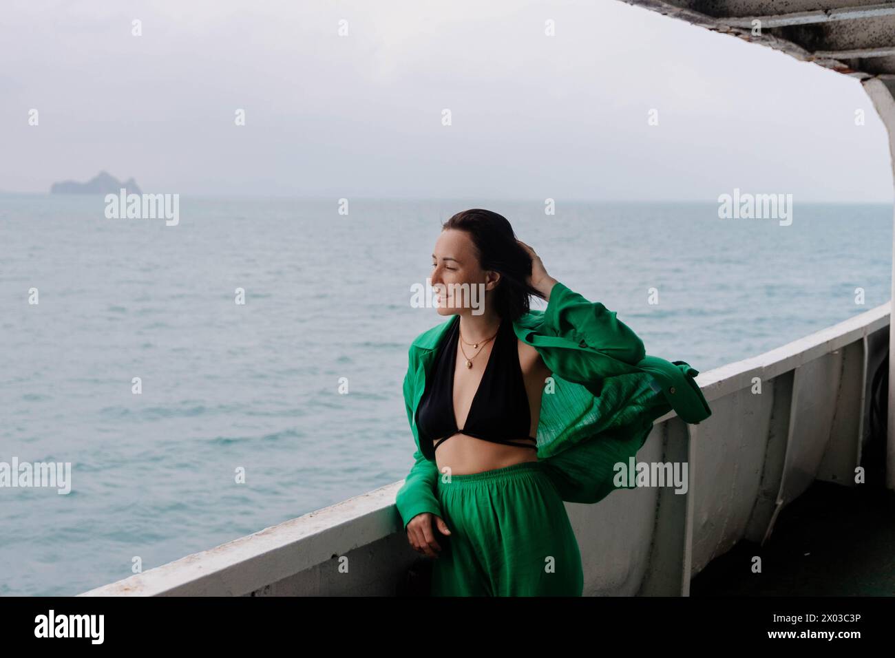 Una donna sorridente si trova su un ponte di traghetto, i suoi capelli vengono tormentati dal vento del mare, con cieli nuvolosi e terra lontana Foto Stock