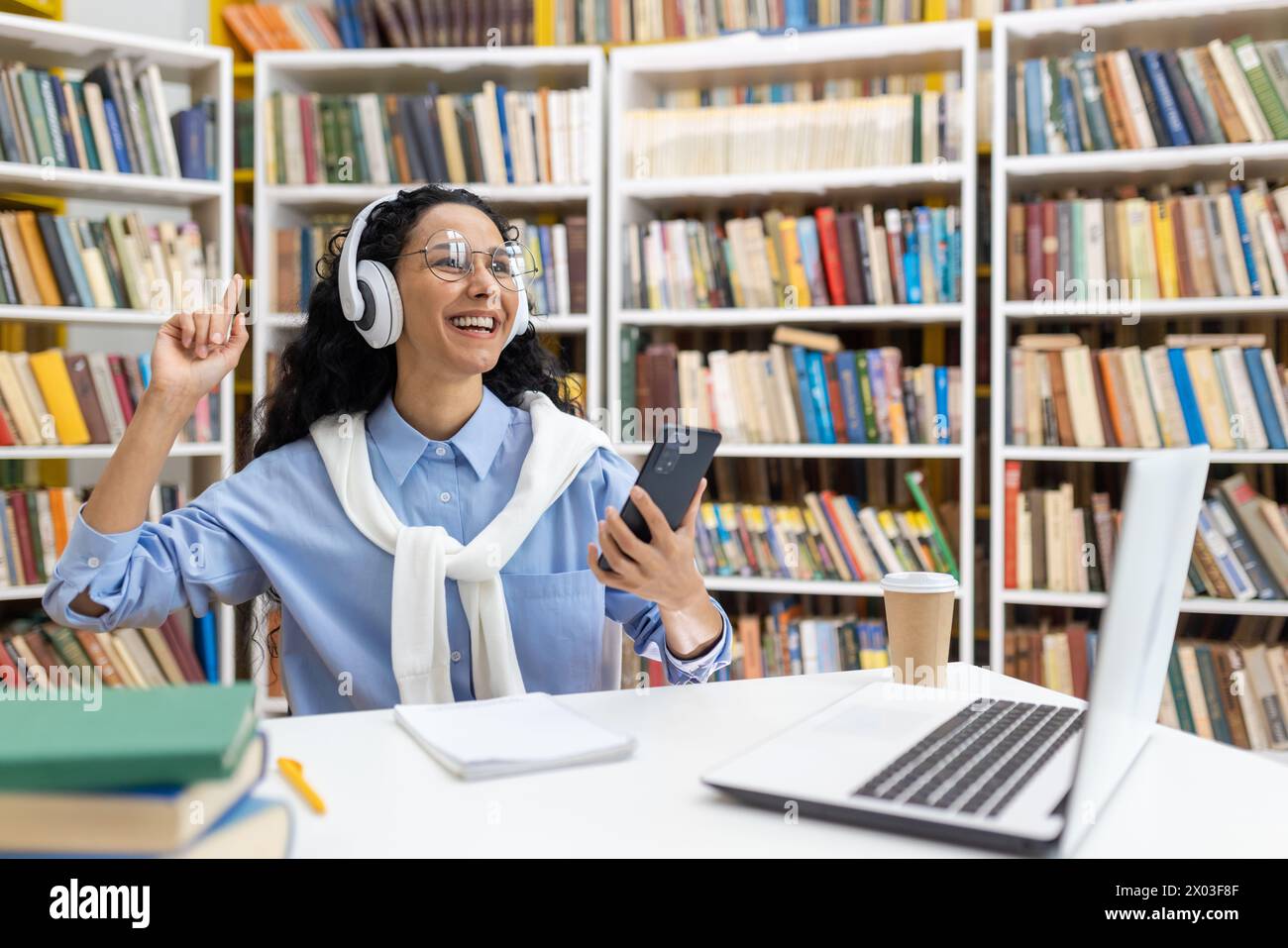 Uno studente allegro sta ascoltando musica con le cuffie, utilizzando uno smartphone in una libreria, circondato da libri e un computer portatile. Foto Stock