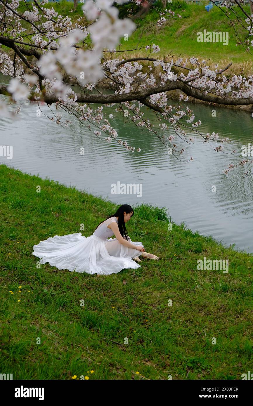 Persona in abito bianco seduta accanto a un fiume calmo sotto i fiori di ciliegio in primavera, catturando un momento tranquillo e riflessivo in Giappone. Foto Stock