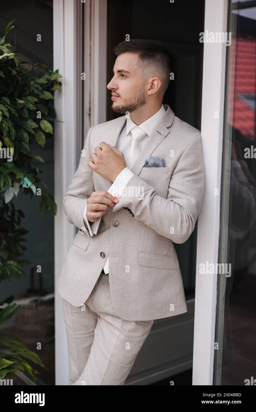 Ritratto di un uomo elegante in terrazza vicino alla finestra e in posa per il fotografo. Bellissimo cresciuto in un abito di colore chiaro Foto Stock